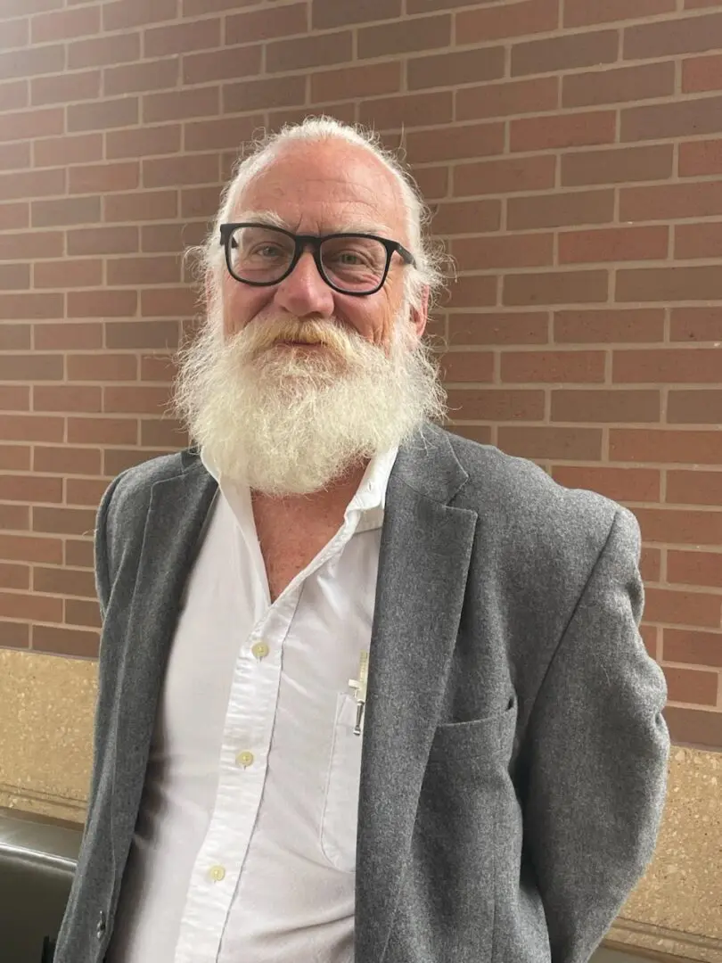 A man with white beard and glasses standing in front of a brick wall.